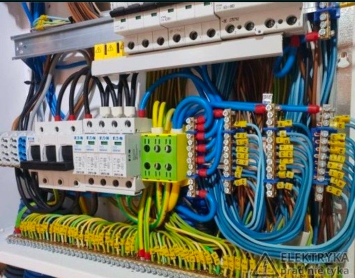 Skomplikowana instalacja elektryczna w rozdzielnicy, widoczne liczne przewody w kolorach niebieskim, żółto-zielonym i czarnym, zabezpieczenia nadprądowe, listwy zaciskowe i elementy łączeniowe.