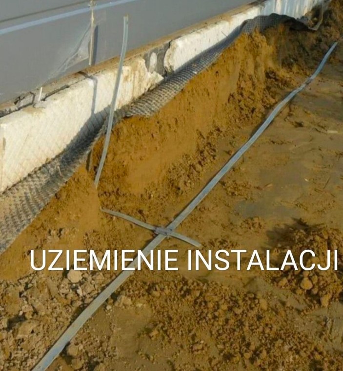 Uziemienie instalacji w wykopie, widoczne metalowe taśmy uziemiające krzyżujące się w ziemi, obok fundament budynku z siatką drenażową.