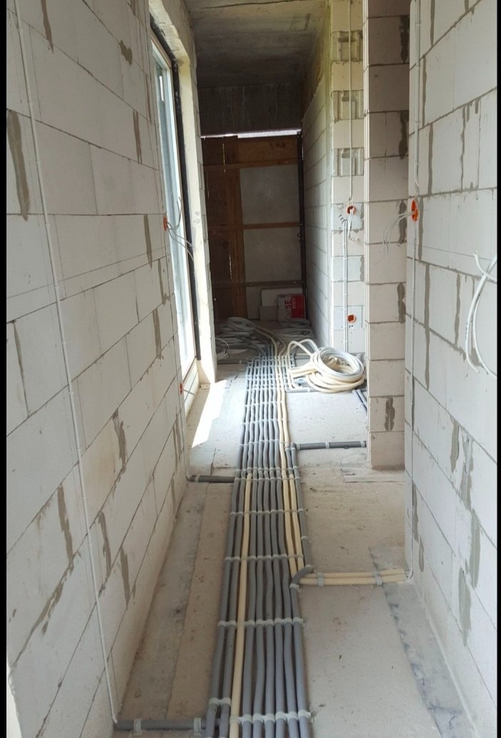 Korytarz w surowym stanie z widocznymi przewodami elektrycznymi ułożonymi w korytkach na podłodze oraz instalacją w ścianach z białego betonu komórkowego. Widoczne puszki instalacyjne.