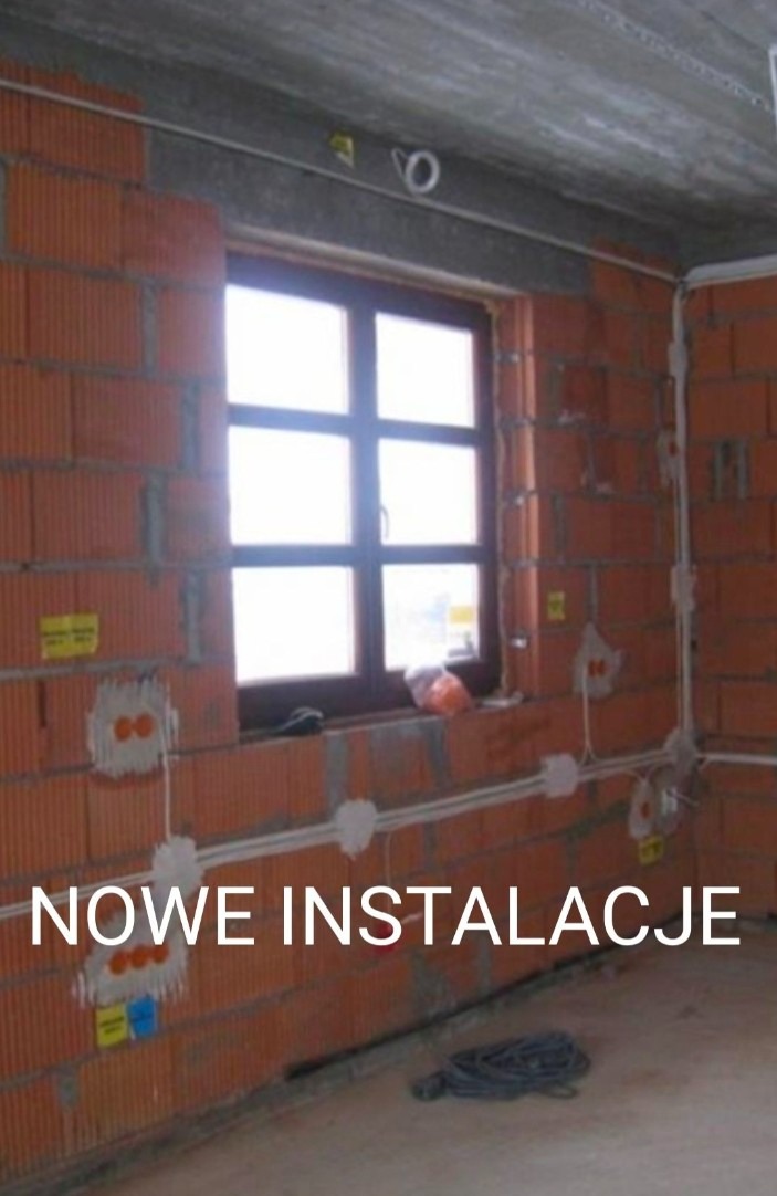 Surowa ściana z cegły ceramicznej z widoczną, nową instalacją elektryczną: białe przewody prowadzone po ścianie, puszki instalacyjne pod oknem, zwój kabla na podłodze.