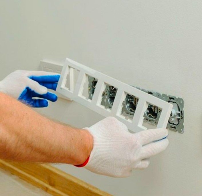 Elektryk w białych rękawicach z niebieskimi palcami montuje ramkę z czterema otworami na gniazdka elektryczne w ścianie.