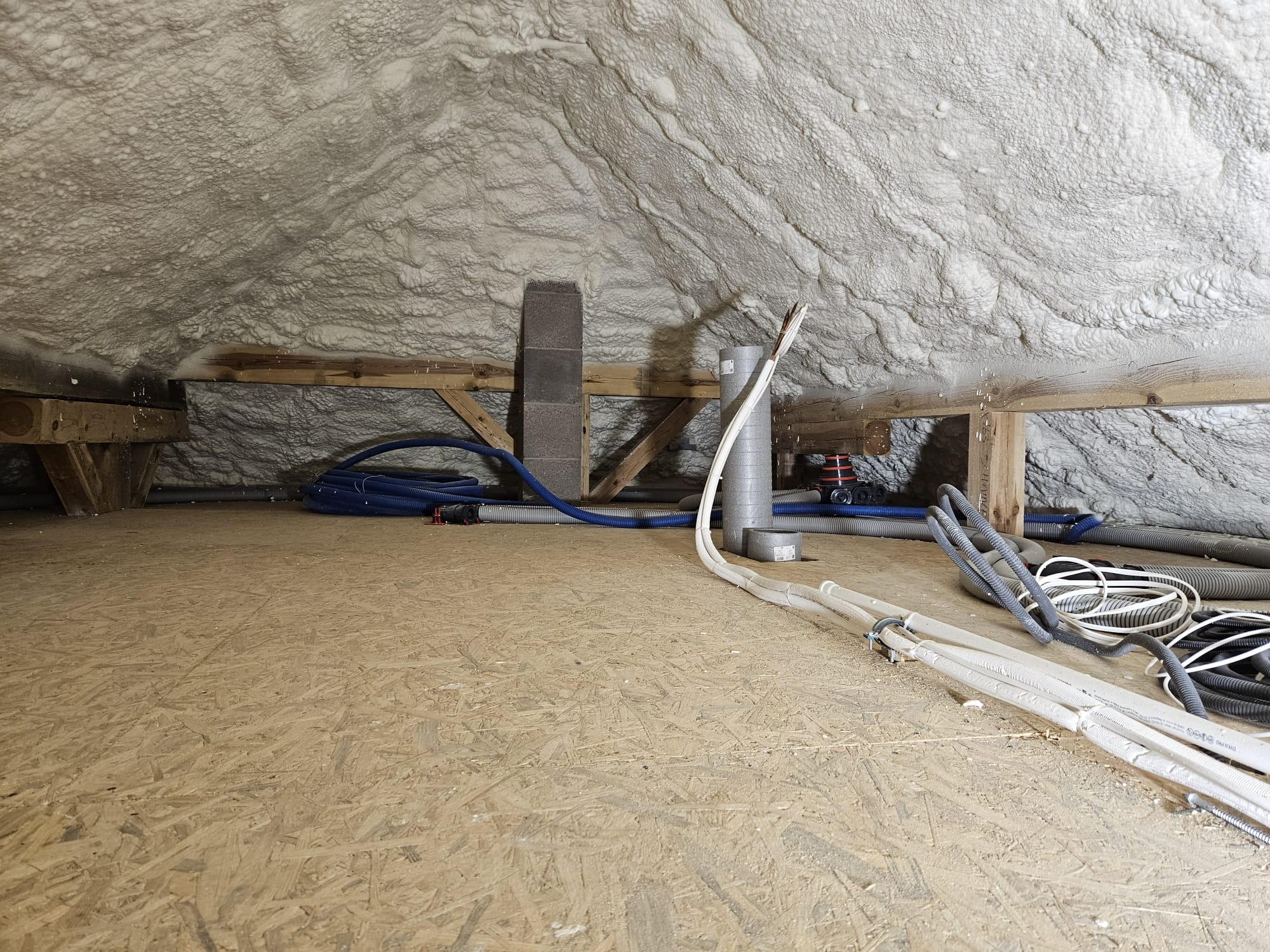 Poddasze po ociepleniu, pokryte grubą warstwą pianki izolacyjnej, widoczne drewniane elementy konstrukcyjne oraz instalacje: rury wentylacyjne, przewody elektryczne i komin z pustaków.