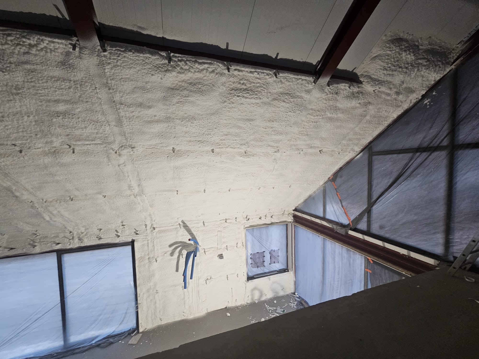 Poddasze z ociepleniem pianką poliuretanową, widoczne okna zabezpieczone folią, instalacja elektryczna i metalowe elementy konstrukcyjne dachu.