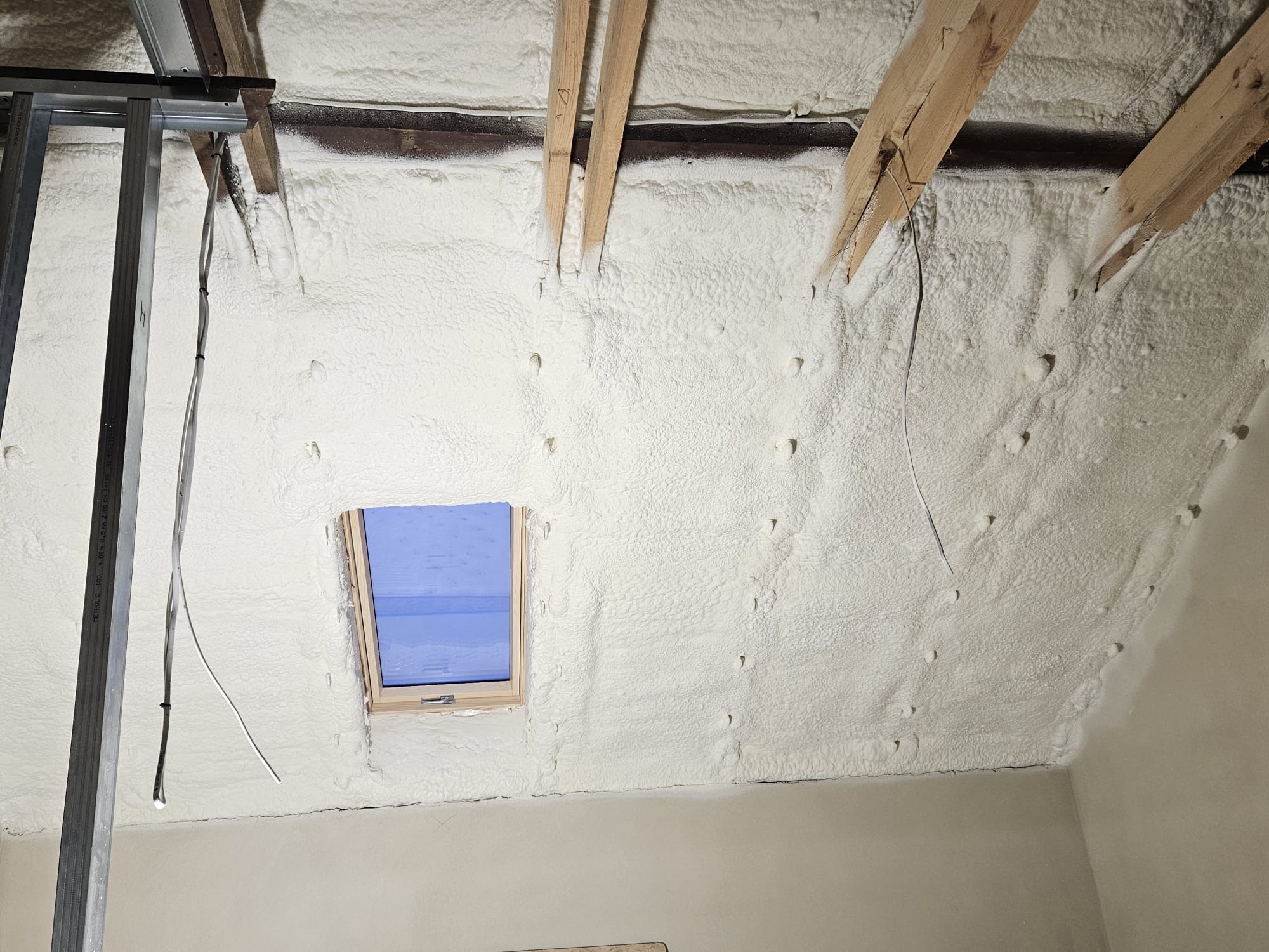 Poddasze z drewnianą konstrukcją i oknem dachowym pokryte grubą warstwą białej pianki izolacyjnej, widoczne metalowe profile konstrukcyjne i wystające przewody.