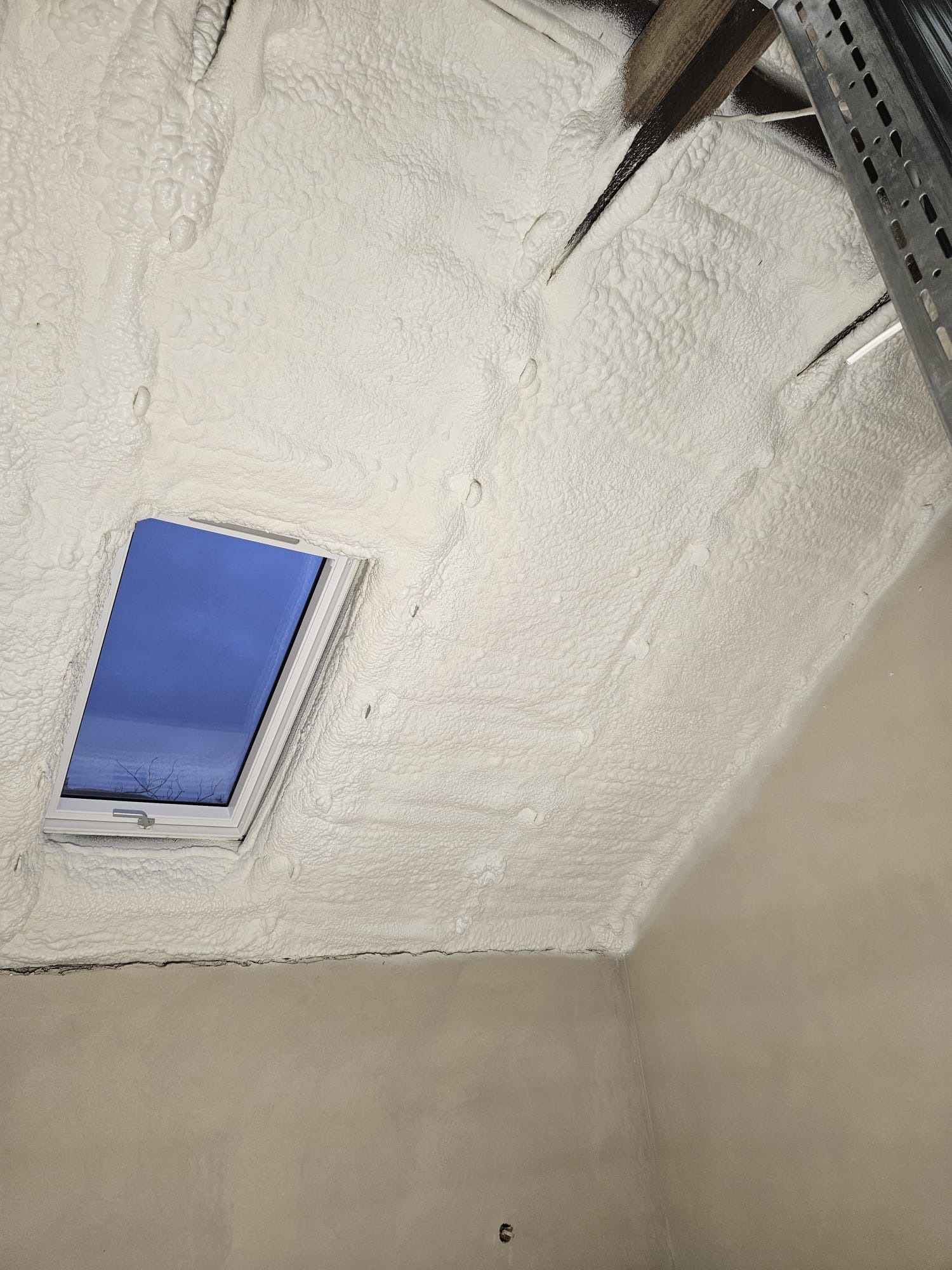 Poddasze z oknem dachowym pokryte grubą warstwą białej pianki izolacyjnej, widoczne fragmenty drewnianej konstrukcji dachu i metalowego stelażu.