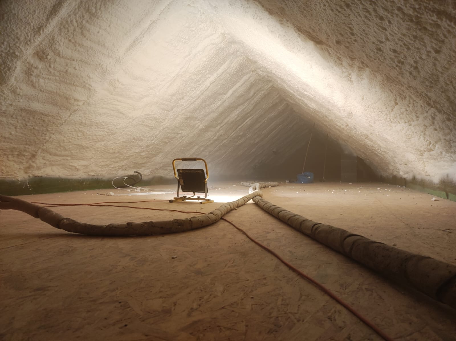 Poddasze po ociepleniu pianką, widoczna struktura pianki, lampa robocza i przewody na drewnianej podłodze.