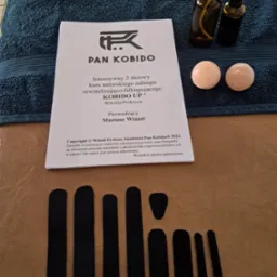 Materiały do kursu Kobido UP: książka z opisem zabiegu, dwie butelki z atomizerem, kamienne kulki do masażu i czarne plastry na beżowym tle.