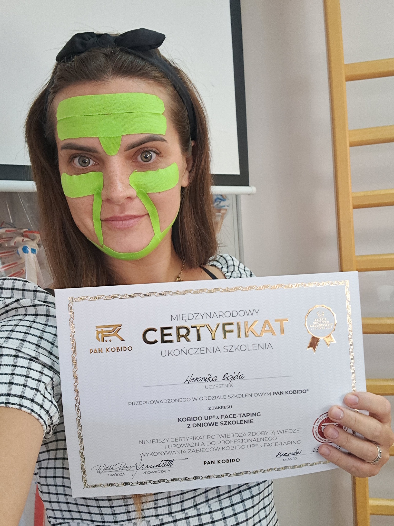 Kobieta z zielonymi taśmami kinesiotaping na twarzy, trzymająca certyfikat ukończenia szkolenia Kobido UP & Face-Taping.