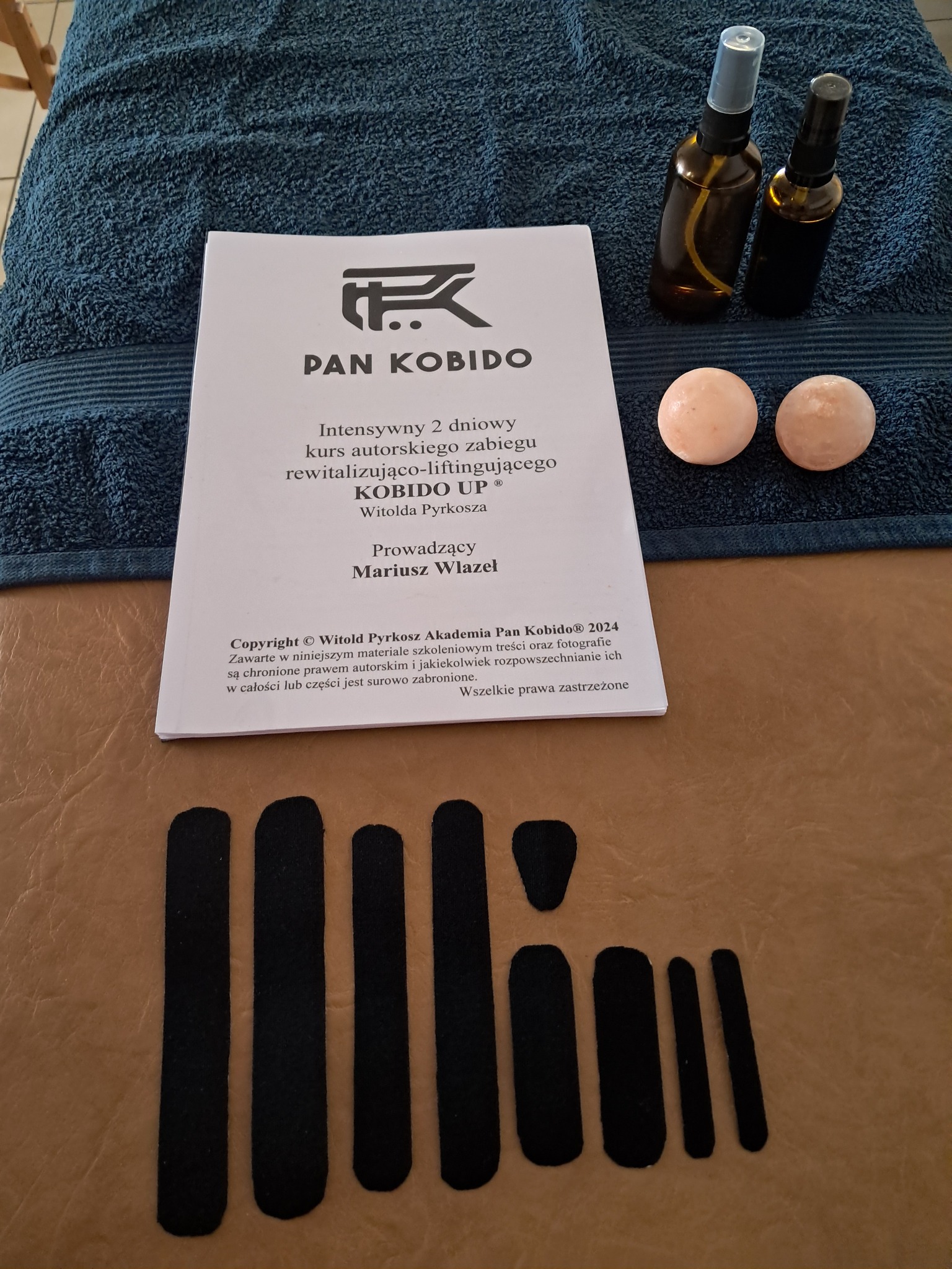 Materiały do kursu Kobido UP: książka z opisem zabiegu, dwie butelki z atomizerem, kamienne kulki do masażu i czarne plastry na beżowym tle.