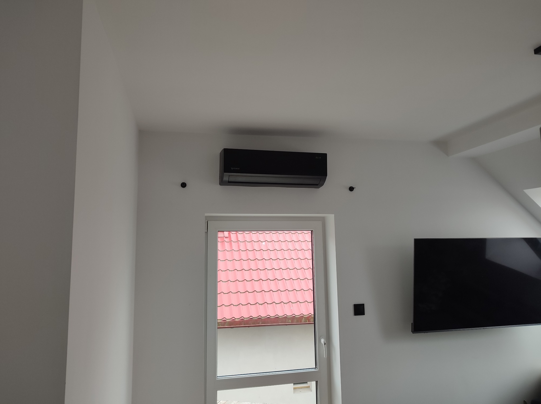 Czarny klimatyzator ścienny zamontowany nad oknem z widokiem na czerwony dach, obok telewizor na białej ścianie.