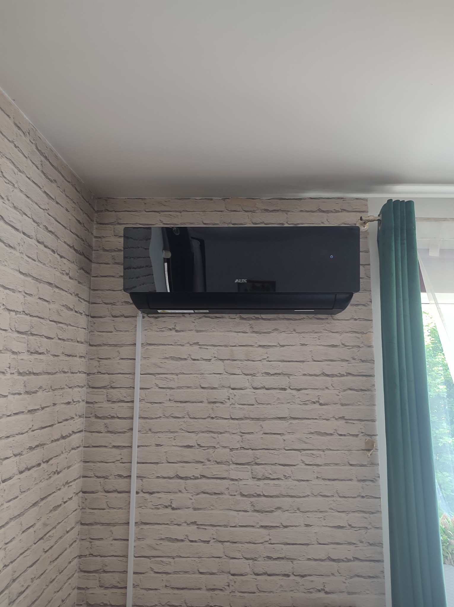 Czarny klimatyzator marki AUX zamontowany na ścianie z imitacją cegły, widoczny fragment zielonej zasłony i biała listwa przypodłogowa.