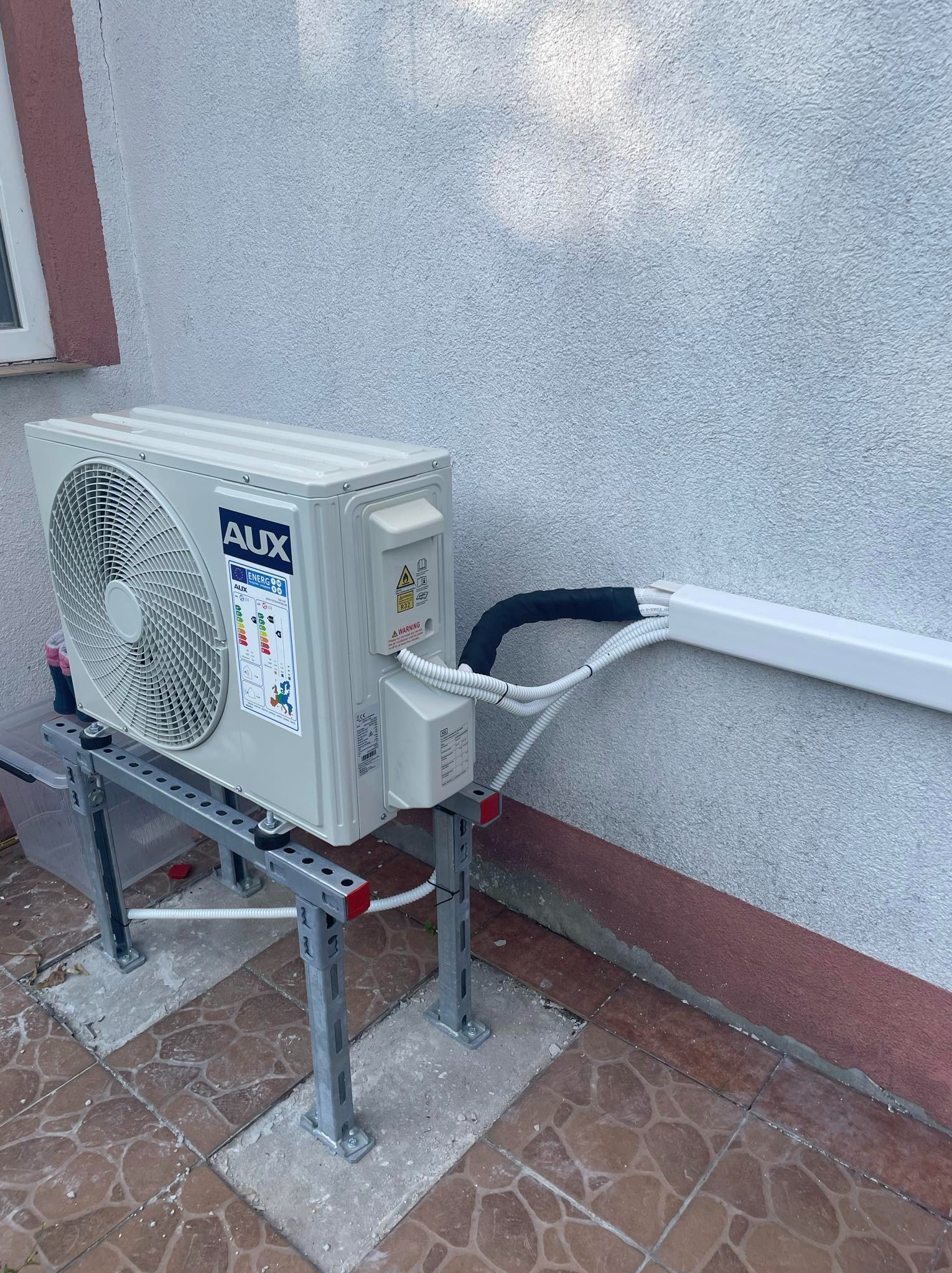 Zewnętrzna jednostka klimatyzacji marki AUX zamontowana na metalowej konstrukcji wsporczej, z widocznymi przewodami w białej listwie, na tle szarej ściany budynku z brązowym cokołem i brązową...