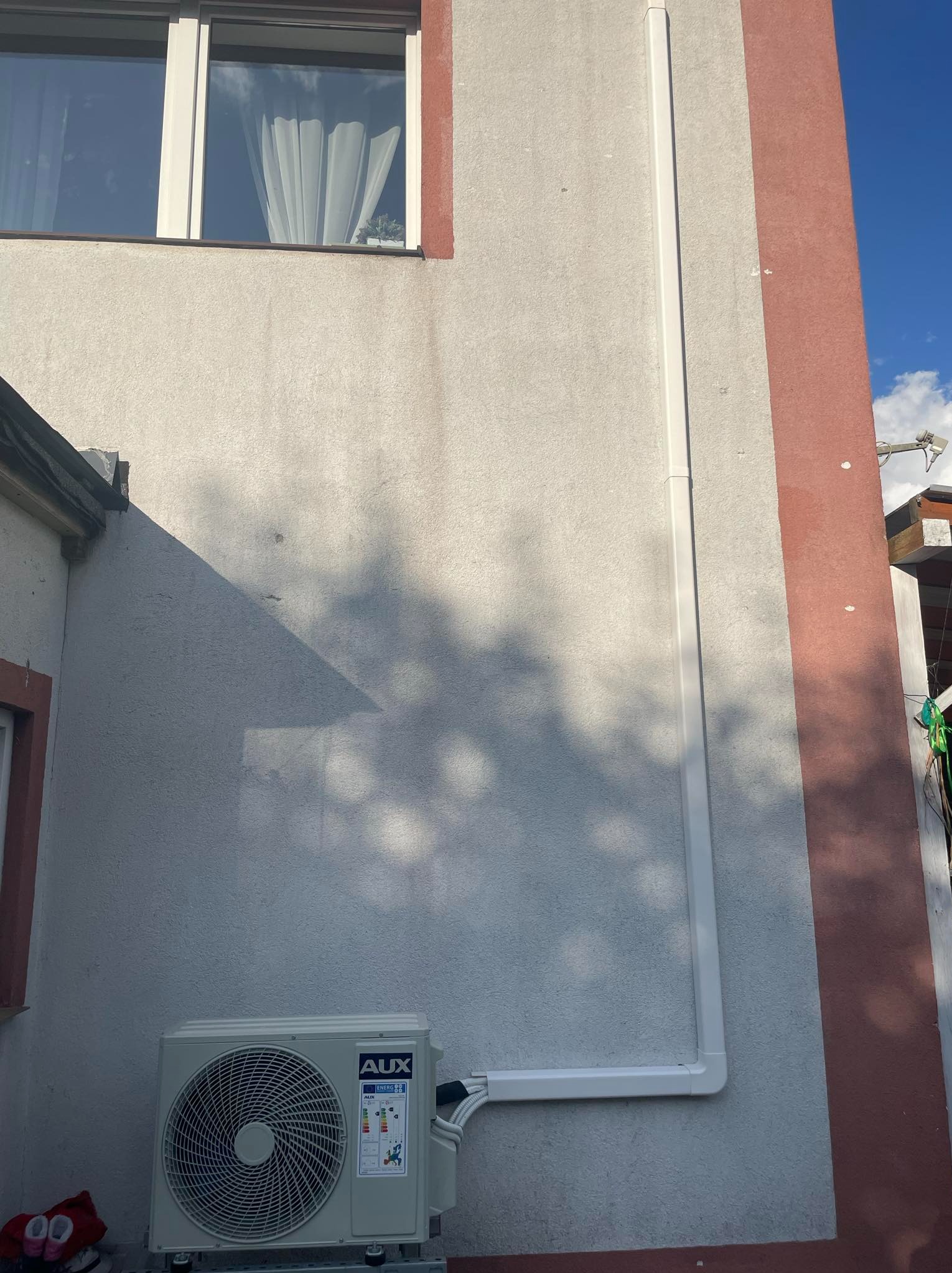 Zewnętrzna jednostka klimatyzacji AUX zamontowana na elewacji budynku w Czersku, z widocznymi rurami odprowadzającymi, cień drzewa na ścianie.