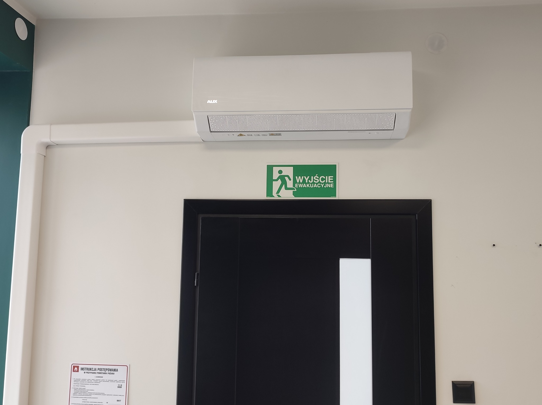 Biały klimatyzator marki AUX zamontowany nad czarnymi drzwiami z zielonym znakiem ewakuacyjnym i instrukcją postępowania w przypadku pożaru na ścianie.