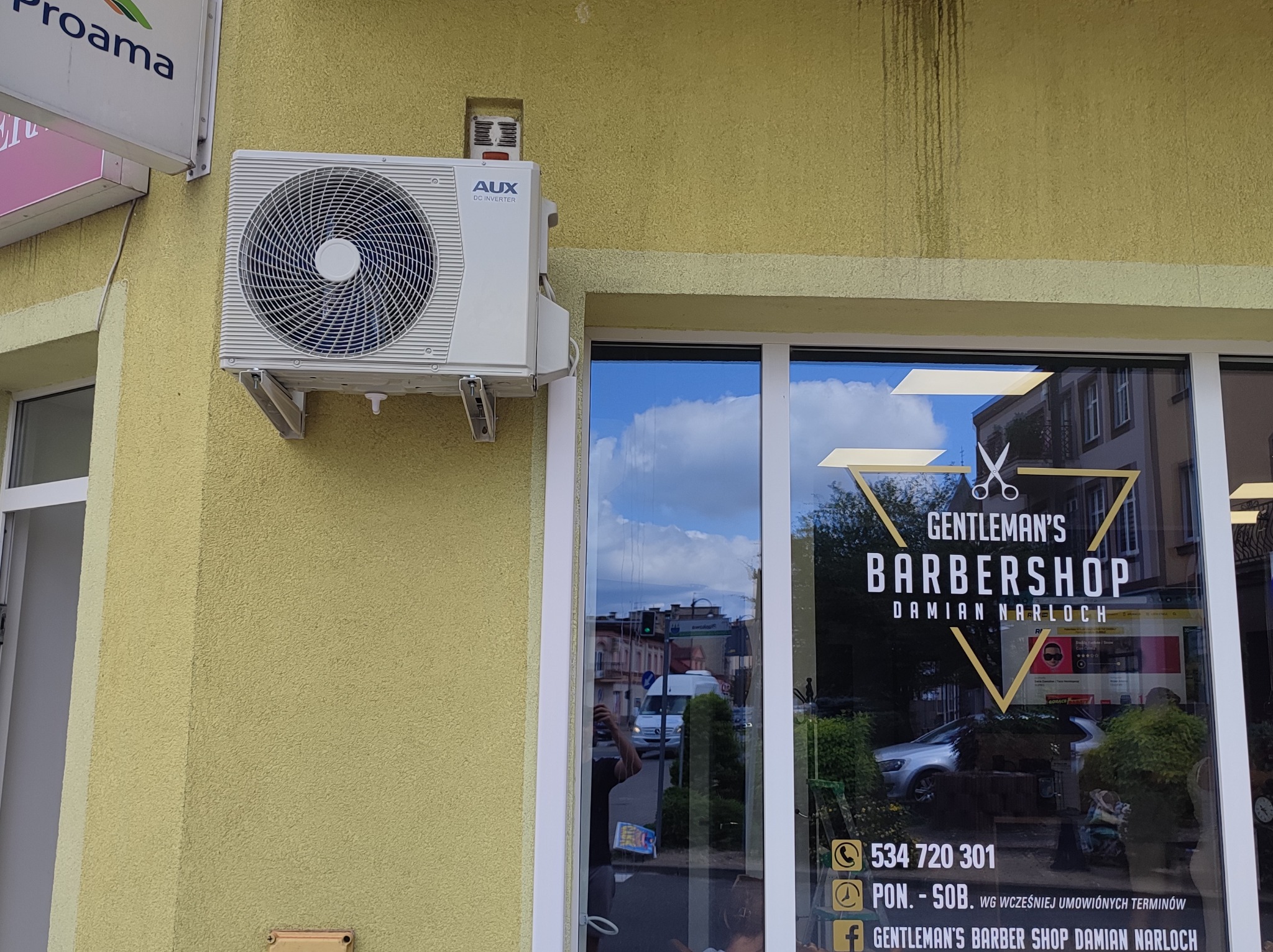 Zewnętrzna jednostka klimatyzacji AUX zamontowana na żółtej, teksturowanej ścianie budynku z widocznym szyldem 'Gentleman's Barbershop' w tle.