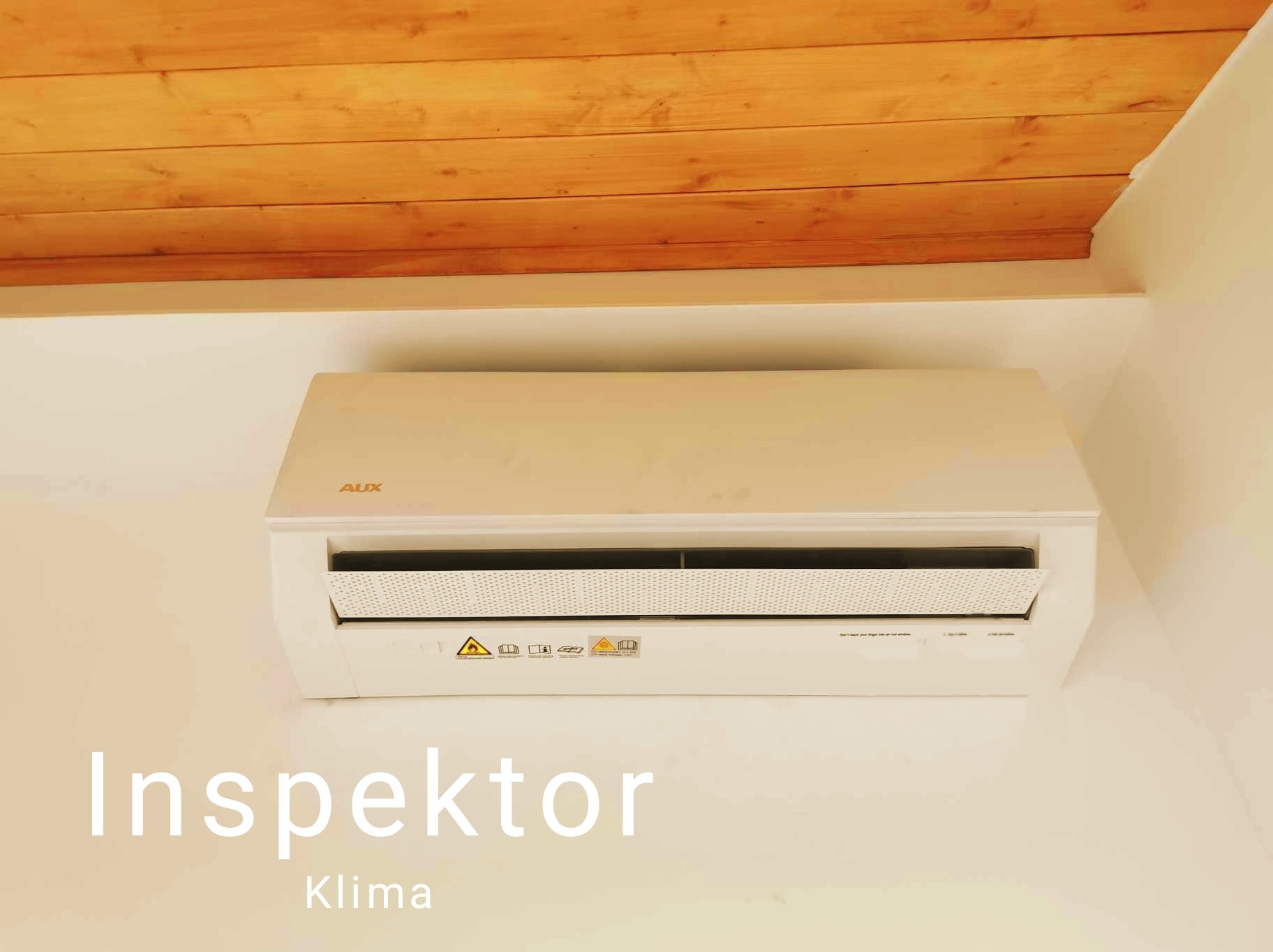 Biała klimatyzacja AUX zamontowana pod drewnianym sufitem na poddaszu, widoczne naklejki ostrzegawcze na urządzeniu.