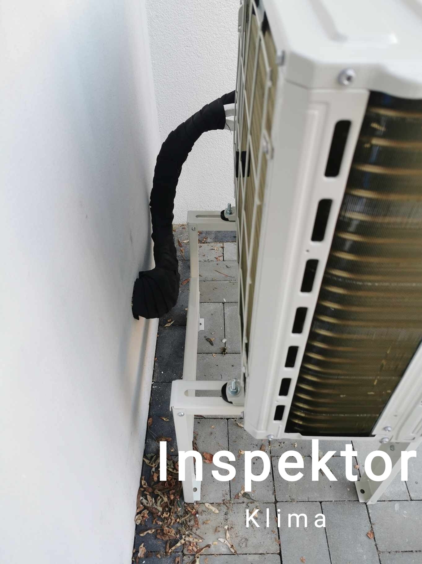 Zewnętrzna jednostka klimatyzacji zamontowana na wsporniku przy białej ścianie budynku w Czersku, widoczna czarna izolacja rury. Kostka brukowa pod spodem.