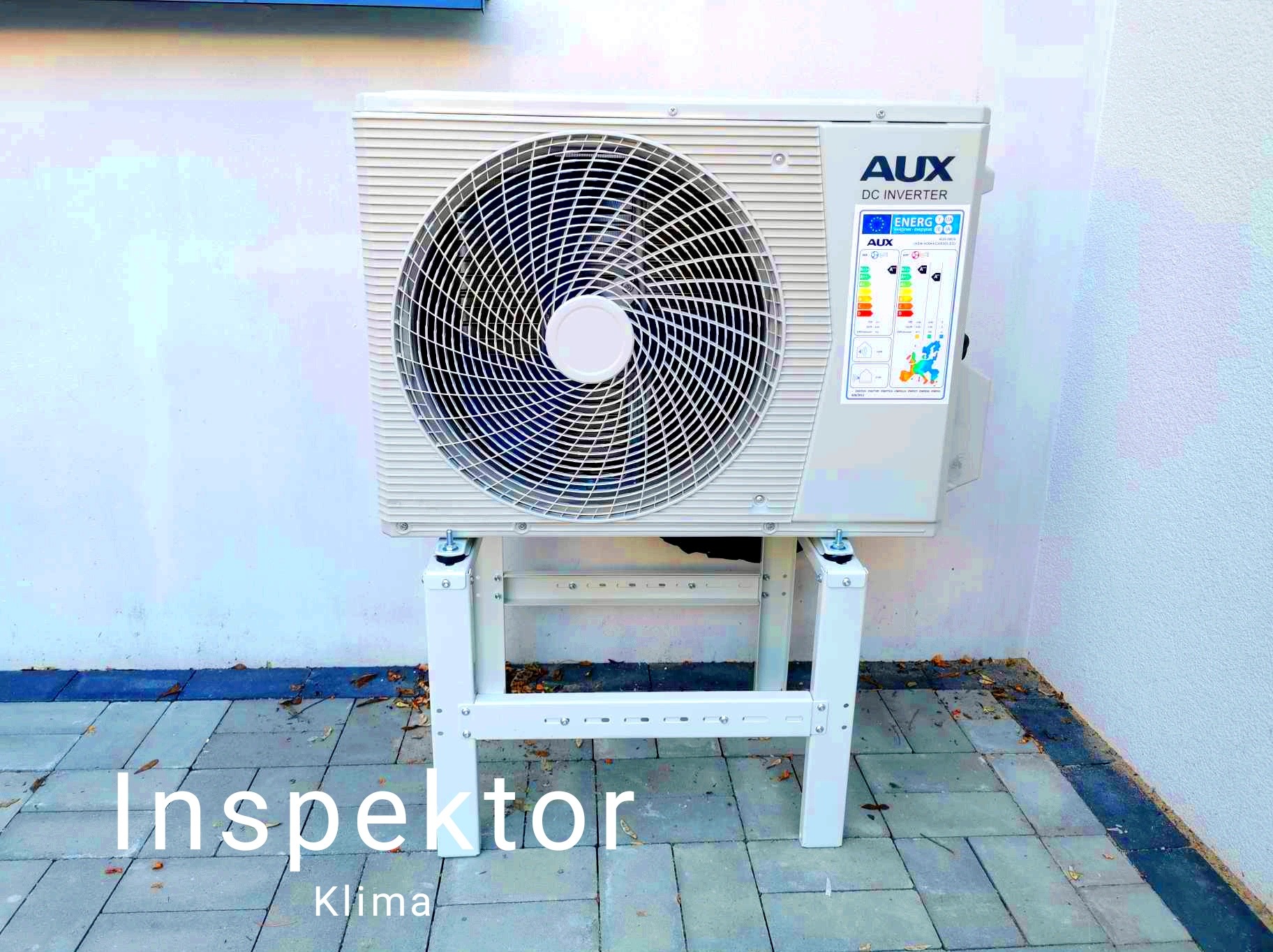 Zewnętrzna jednostka klimatyzacji AUX DC Inverter zamontowana na białym, metalowym stelażu na tle białej ściany i szarej kostki brukowej.