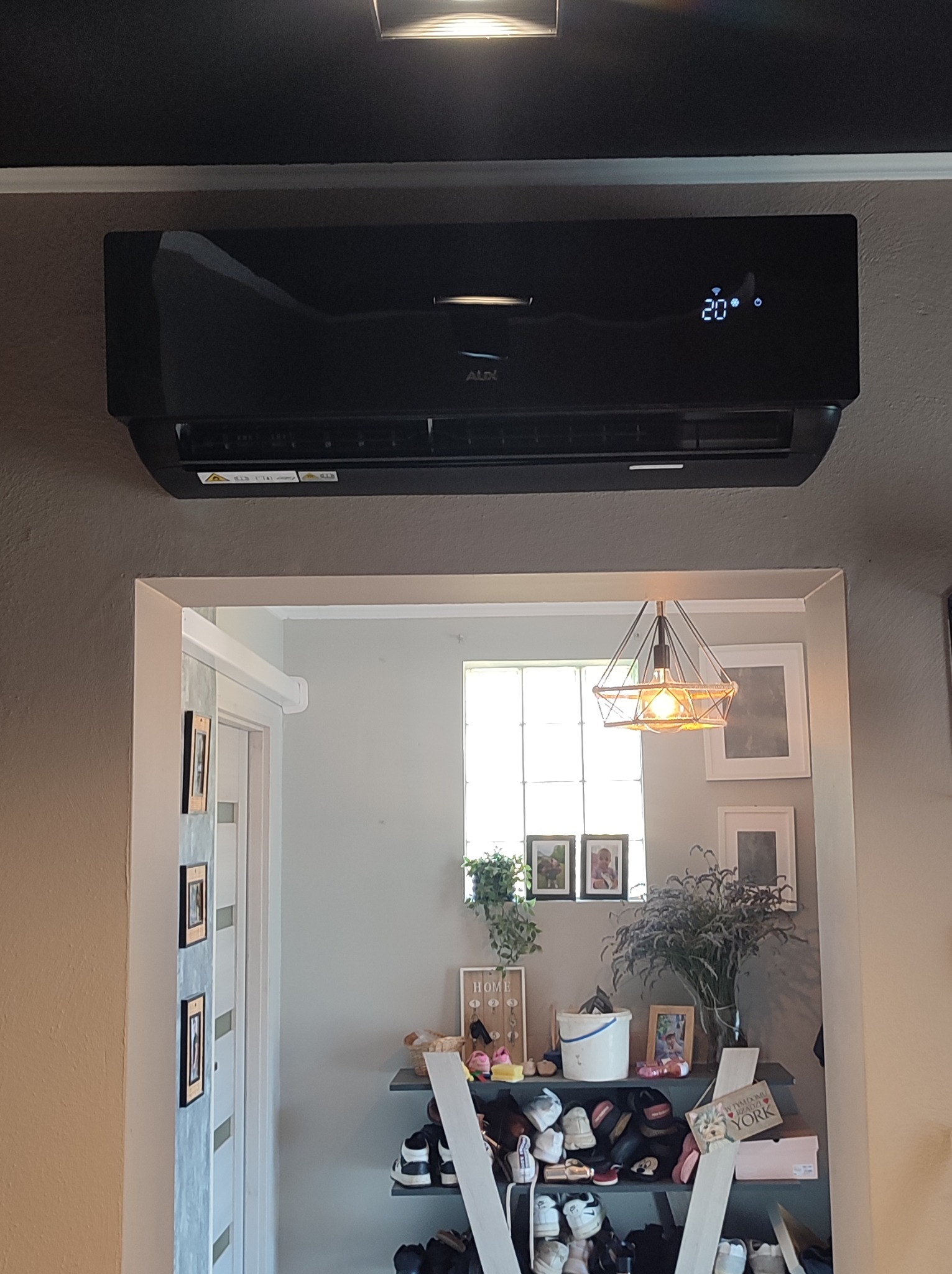 Czarny klimatyzator marki AUX zamontowany na szarej ścianie, widoczny przez futrynę drzwi, z wyświetlaczem temperatury 20 stopni Celsjusza.