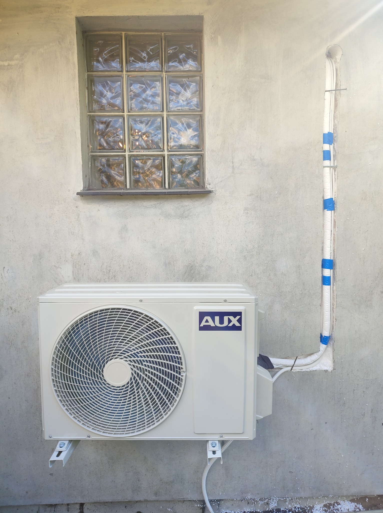 Zewnętrzna jednostka klimatyzacji AUX zamontowana na szarej, chropowatej ścianie budynku, z widoczną instalacją prowadzącą do góry, zabezpieczoną niebieską taśmą, okno z luksferów w tle.