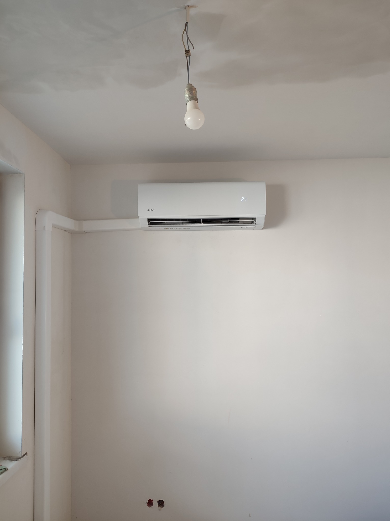 Biały klimatyzator AUX zamontowany na ścianie w jasnym pomieszczeniu, widoczne białe rury instalacyjne i surowa żarówka wisząca z sufitu.