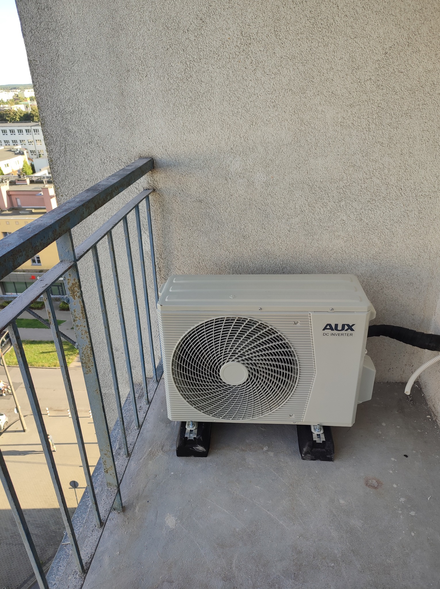 Zewnętrzna jednostka klimatyzacji AUX DC Inverter zamontowana na balkonie, widok z góry, z fragmentem metalowej balustrady i elewacją budynku w tle.
