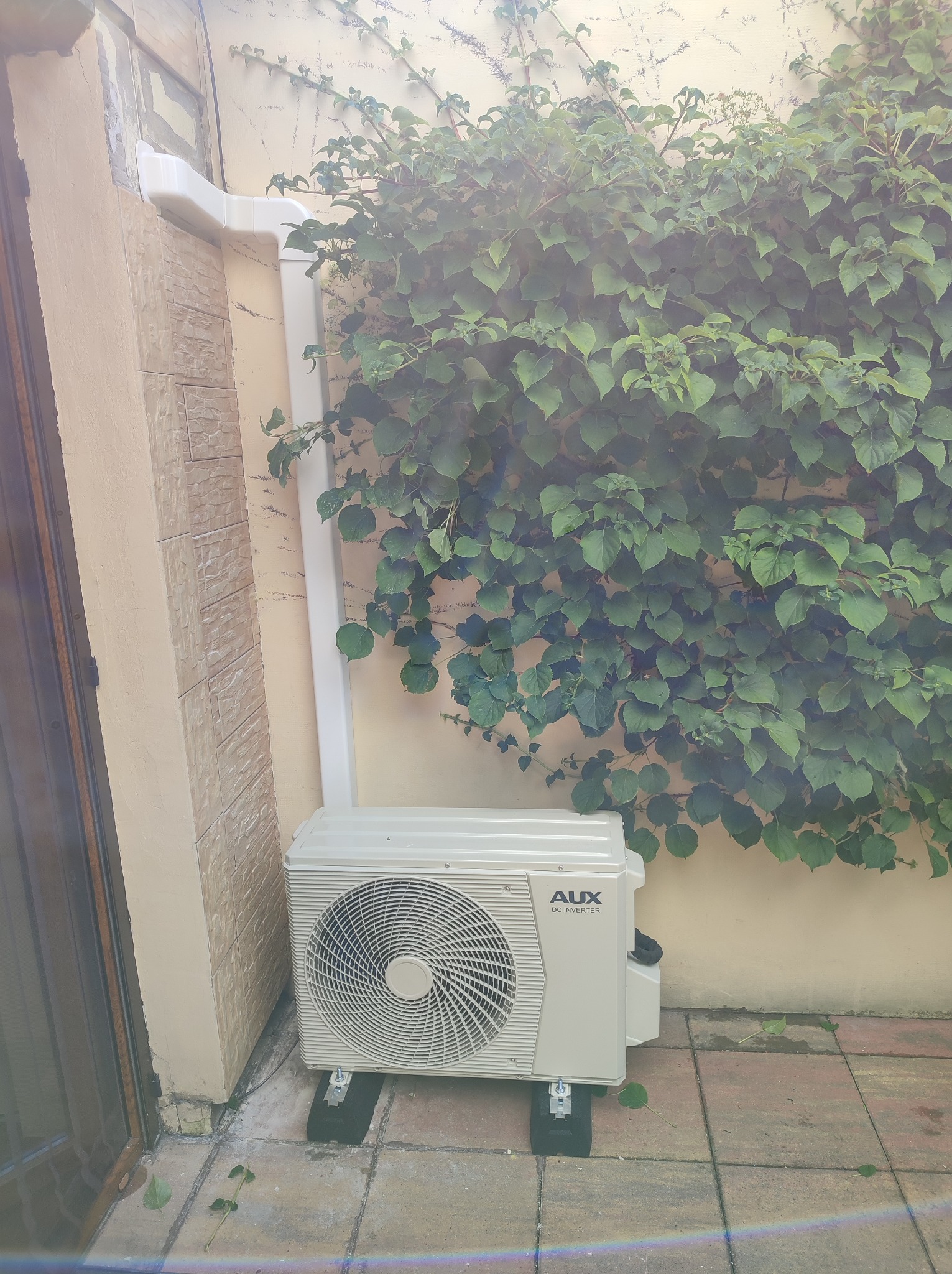 Zewnętrzna jednostka klimatyzacji AUX DC Inverter zamontowana na wspornikach na patio, z rurami odprowadzającymi wilgoć i pnączami na ścianie w tle.