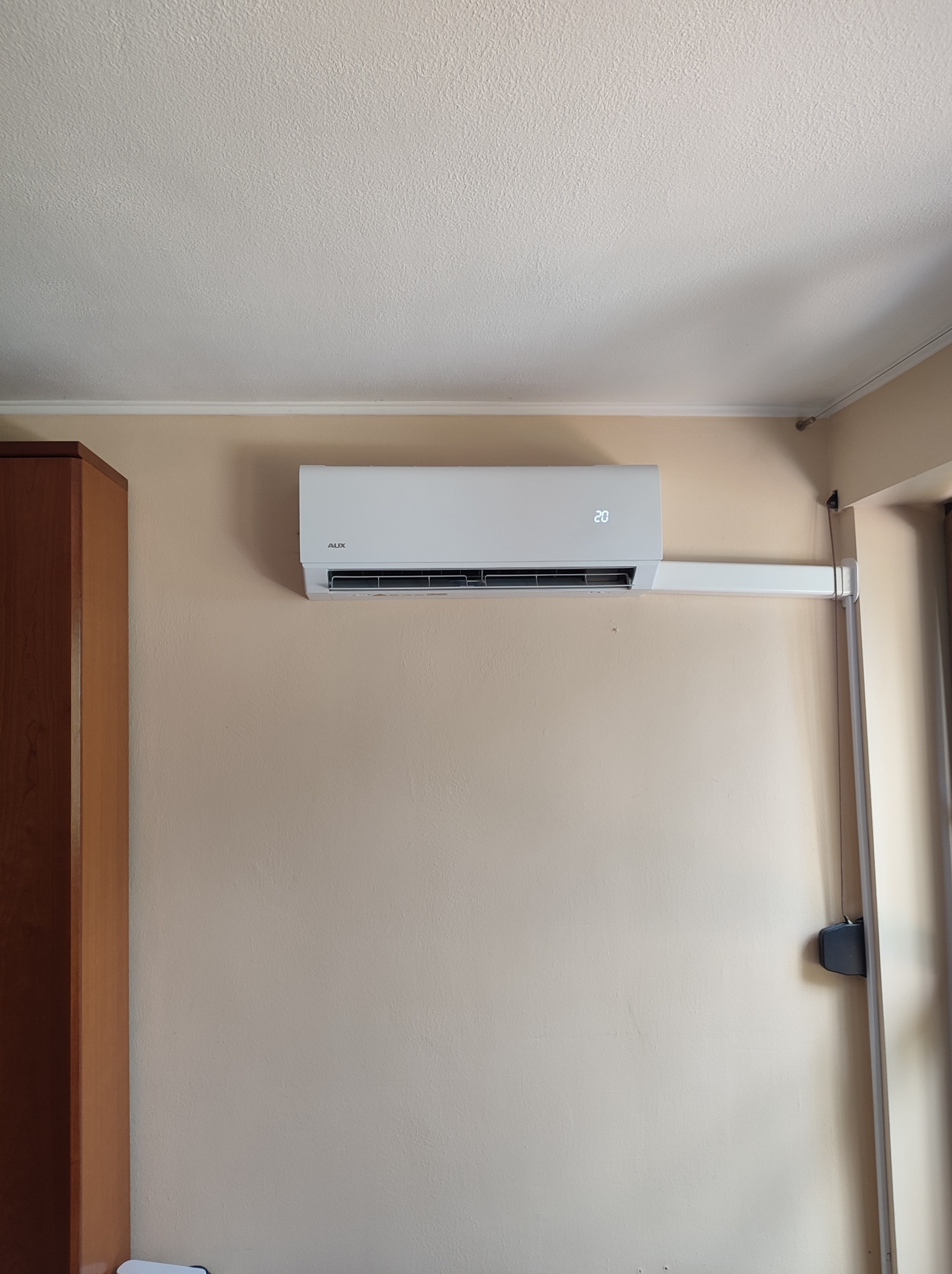 Biała klimatyzacja AUX zamontowana na ścianie w pokoju, widoczny wyświetlacz z temperaturą 20 stopni Celsjusza, obok szafa.