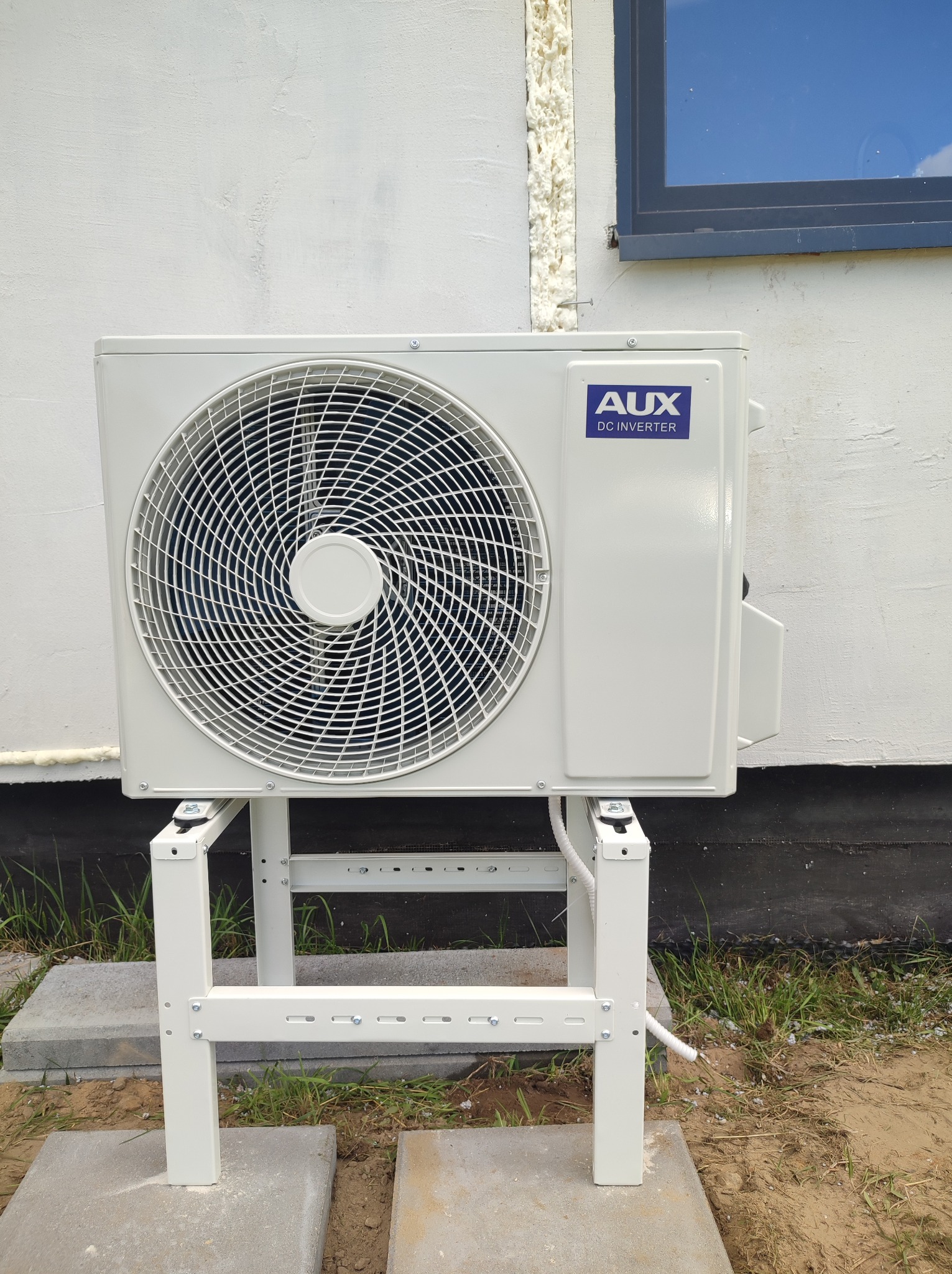 Biała jednostka zewnętrzna klimatyzacji AUX DC Inverter zamontowana na metalowym stelażu na betonowych płytach przy ścianie budynku z widocznym oknem i pianką uszczelniającą.