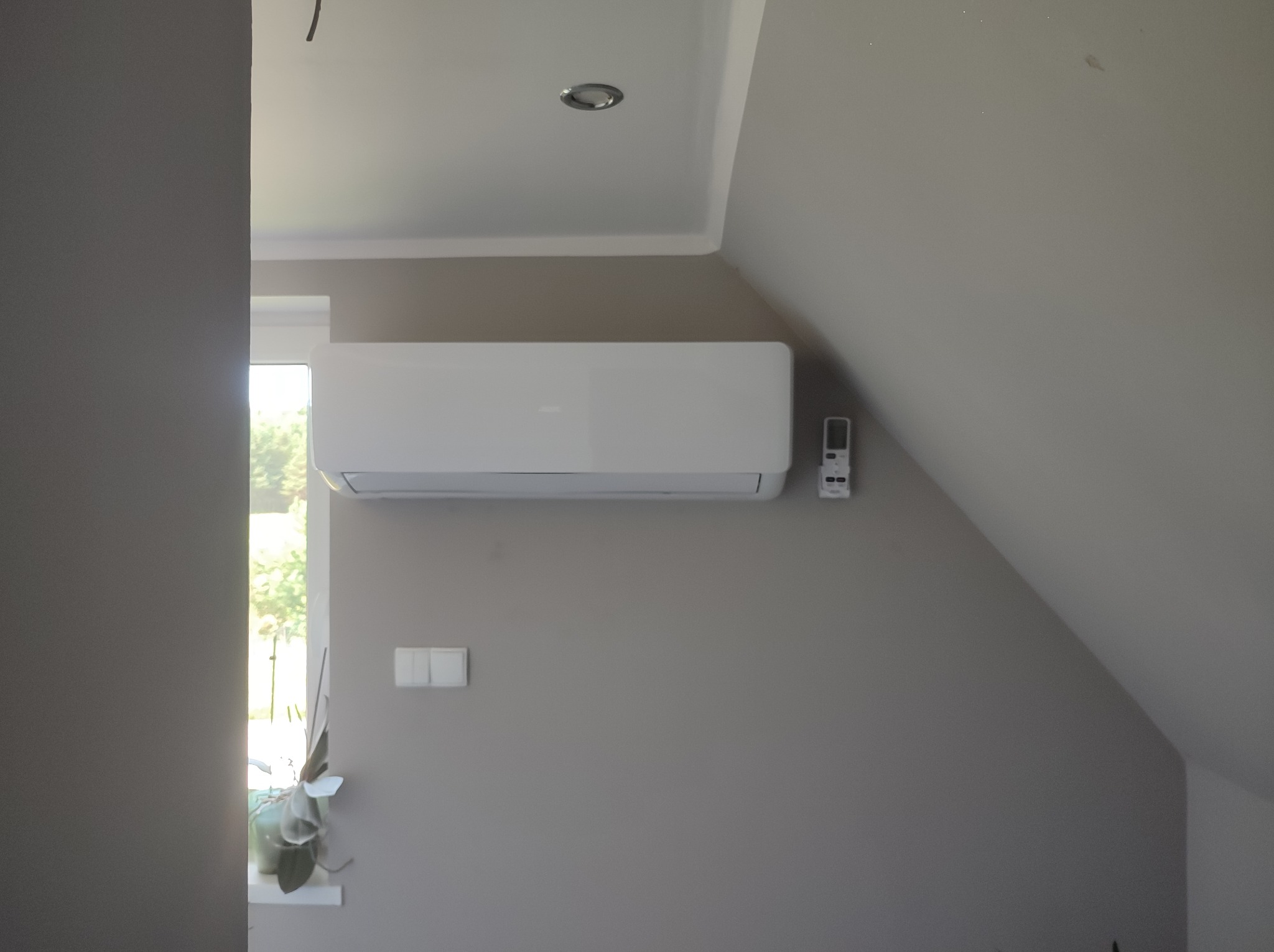Biały klimatyzator ścienny w szarym pokoju na poddaszu, z pilotem zamontowanym obok i rośliną w doniczce przy oknie.