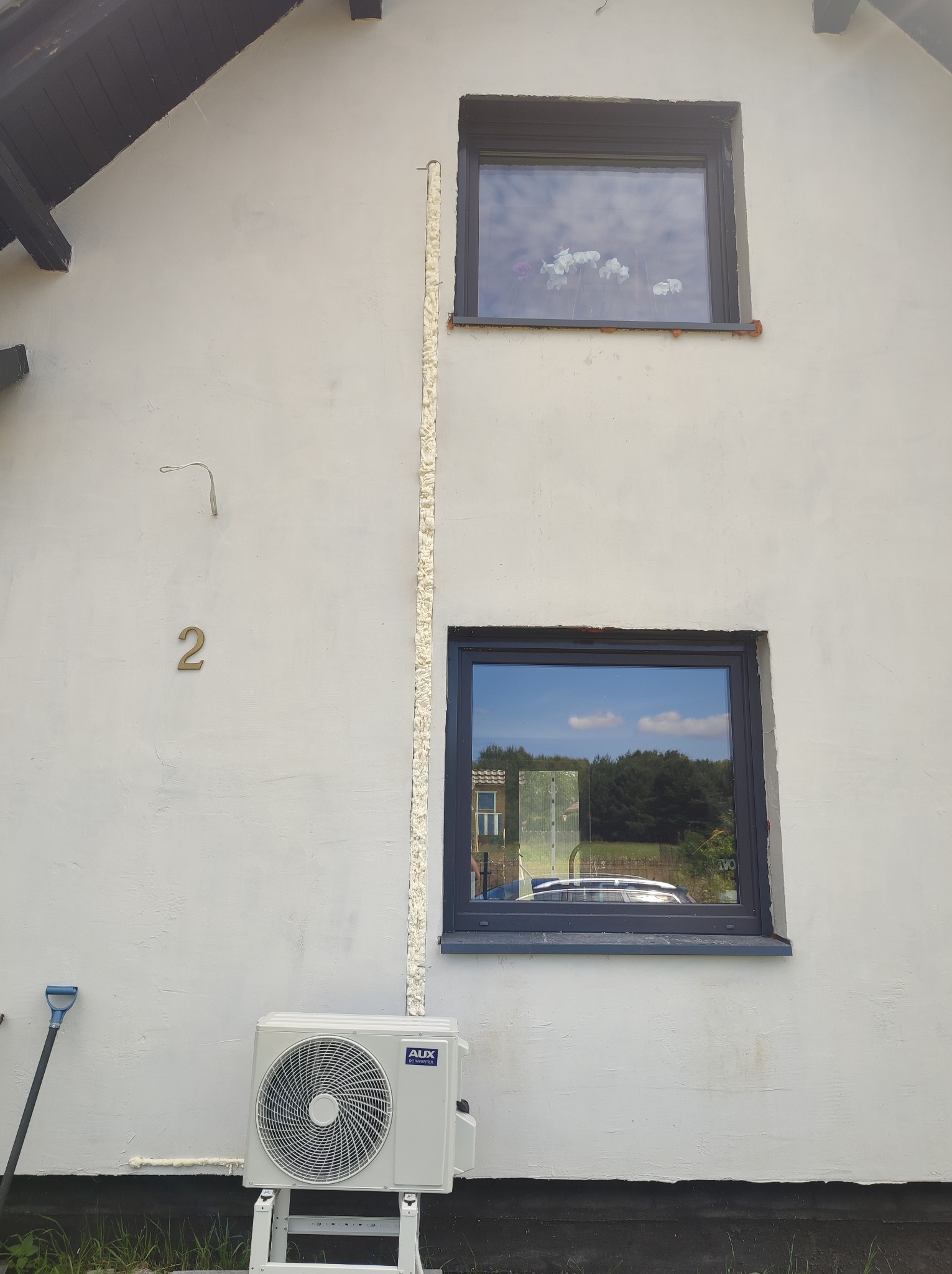 Biała fasada domu z zamontowaną jednostką zewnętrzną klimatyzacji AUX, widoczne okna z ciemnymi ramami i numer domu '2', ślady pianki montażowej.