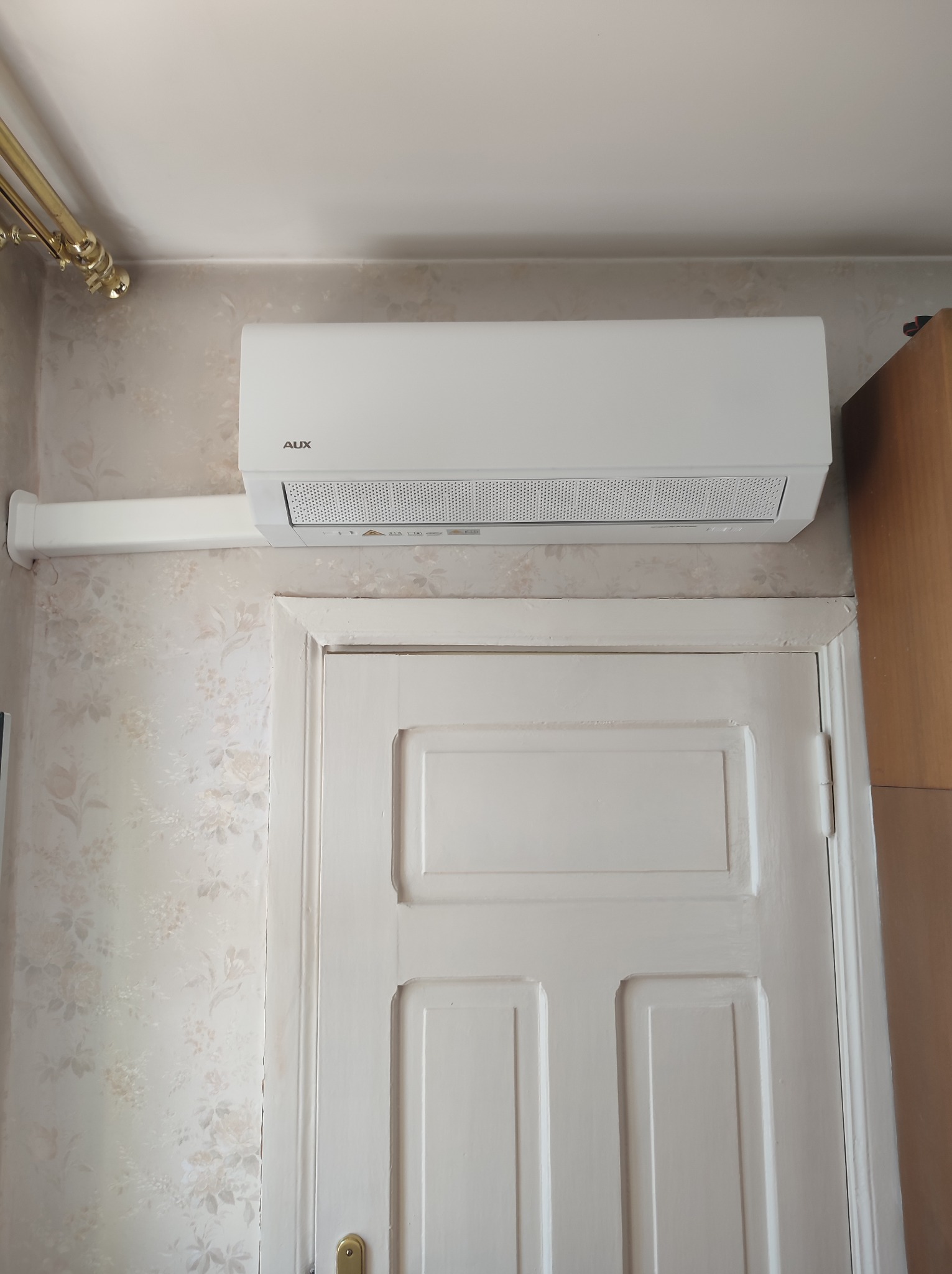 Biała jednostka klimatyzacyjna marki AUX zamontowana nad białą, panelową ścianą i drzwiami w pomieszczeniu z tapetą w kwiaty.