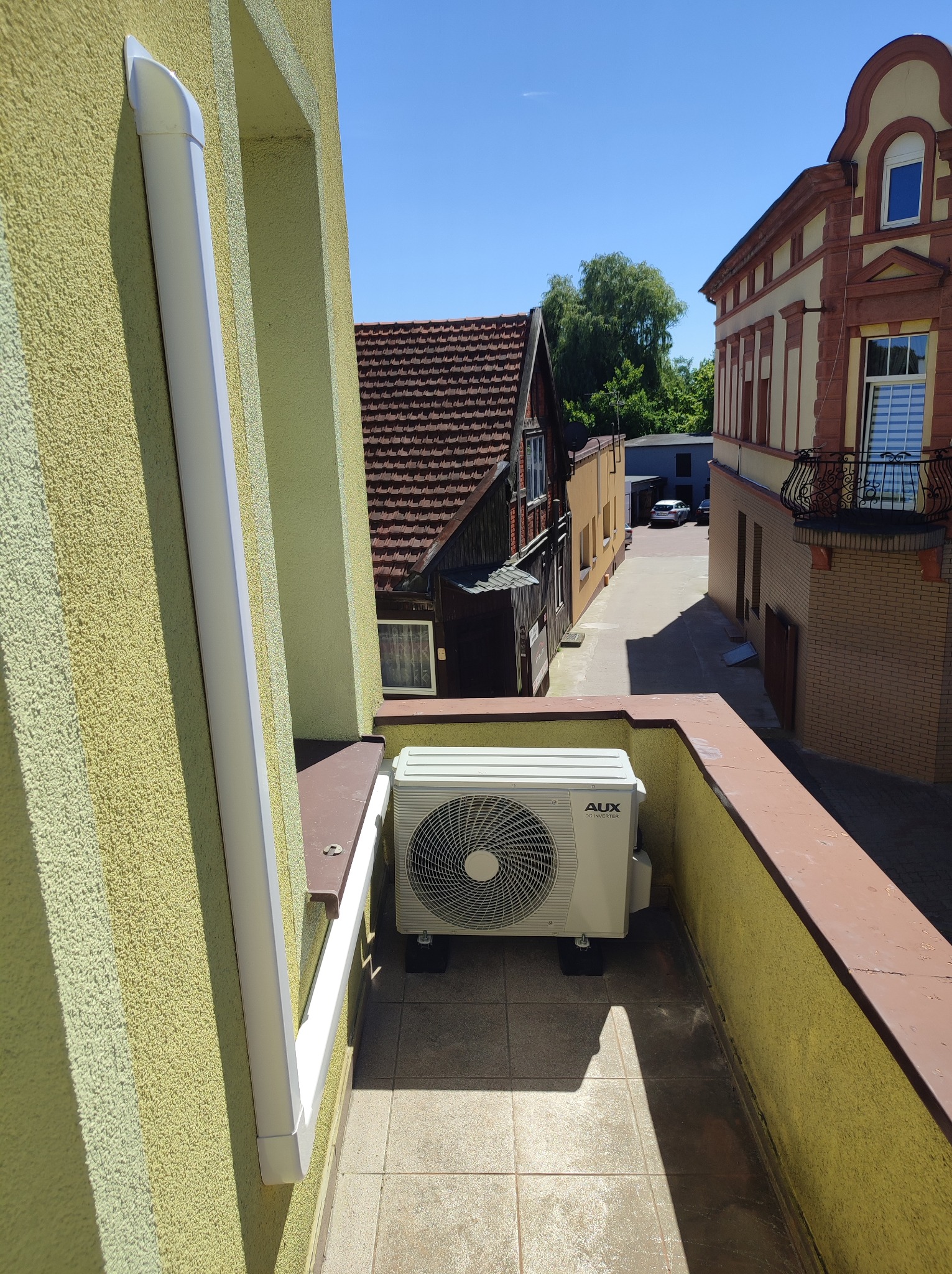Jednostka zewnętrzna klimatyzacji AUX DC Inverter zamontowana na balkonie budynku mieszkalnego, widok z góry na ulicę z zabudową kamieniczną w tle.