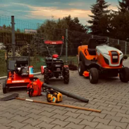 Zestaw sprzętu ogrodniczego: aerator Weibang, glebogryzarka, kosiarka traktorek Stihl, piła spalinowa, dmuchawa do liści i grabie na kostce brukowej, widok na ogrodzenie i zachmurzone niebo.