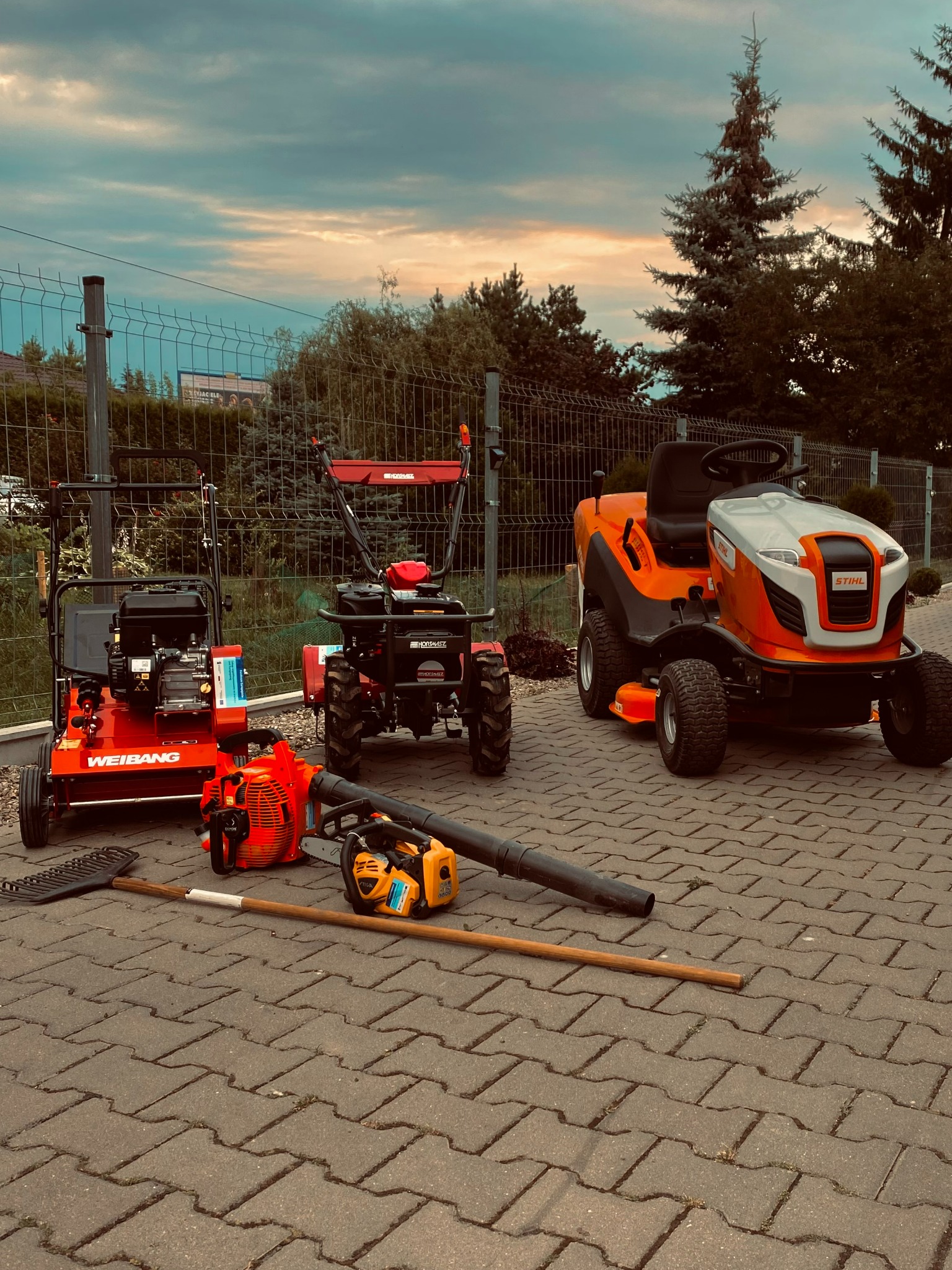 Zestaw sprzętu ogrodniczego: aerator Weibang, glebogryzarka, kosiarka traktorek Stihl, piła spalinowa, dmuchawa do liści i grabie na kostce brukowej, widok na ogrodzenie i zachmurzone niebo.