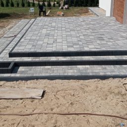 Monika Niewazne - Ułożony taras z kostki brukowej z trzema stopniami, wykończony ciemnoszarym obrzeżem, widoczny piasek i narzędzia brukarskie w tle.