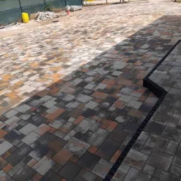 Ułożona nawierzchnia z wielokolorowej kostki brukowej w odcieniach szarości, brązu i beżu, oddzielona czarnym obrzeżem, z widocznym placem budowy w tle.