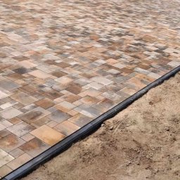 Monika Niewazne - Ułożona nawierzchnia z kostki brukowej w odcieniach brązu i szarości, oddzielona czarnym obrzeżem od piaszczystego podłoża.