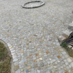 Ułożona z drobnej kostki granitowej nawierzchnia z okrągłym wzorem pośrodku, otoczona zielenią i elementami muru z kamienia.