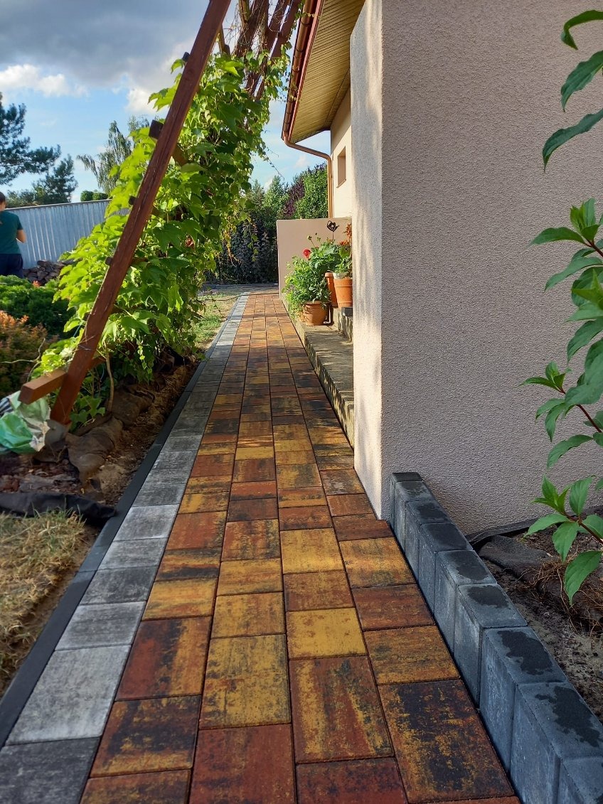 Kolorowa ścieżka z kostki brukowej prowadząca wzdłuż domu, obrzeża z szarej kostki, pergola z pnączami, widok na ogród.