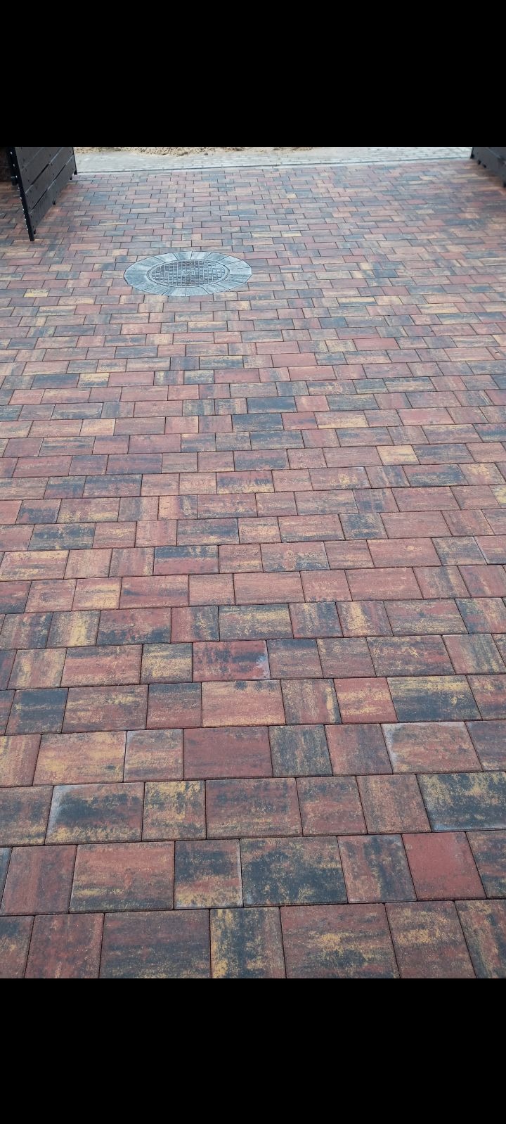 Nowo ułożona, wielobarwna kostka brukowa w odcieniach czerwieni, brązu i czerni, z okrągłą studzienką kanalizacyjną pośrodku, widziana z perspektywy lekko z góry.