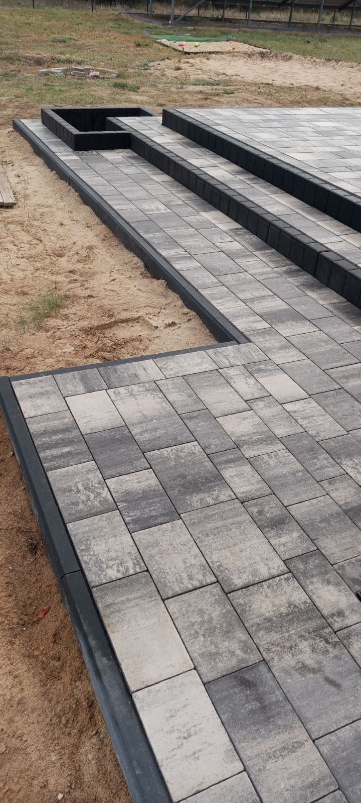 Ułożona, szara kostka brukowa z ciemnoszarym obrzeżem, tworząca taras z podwyższeniem i miejscem na rośliny, otoczona piaskiem.