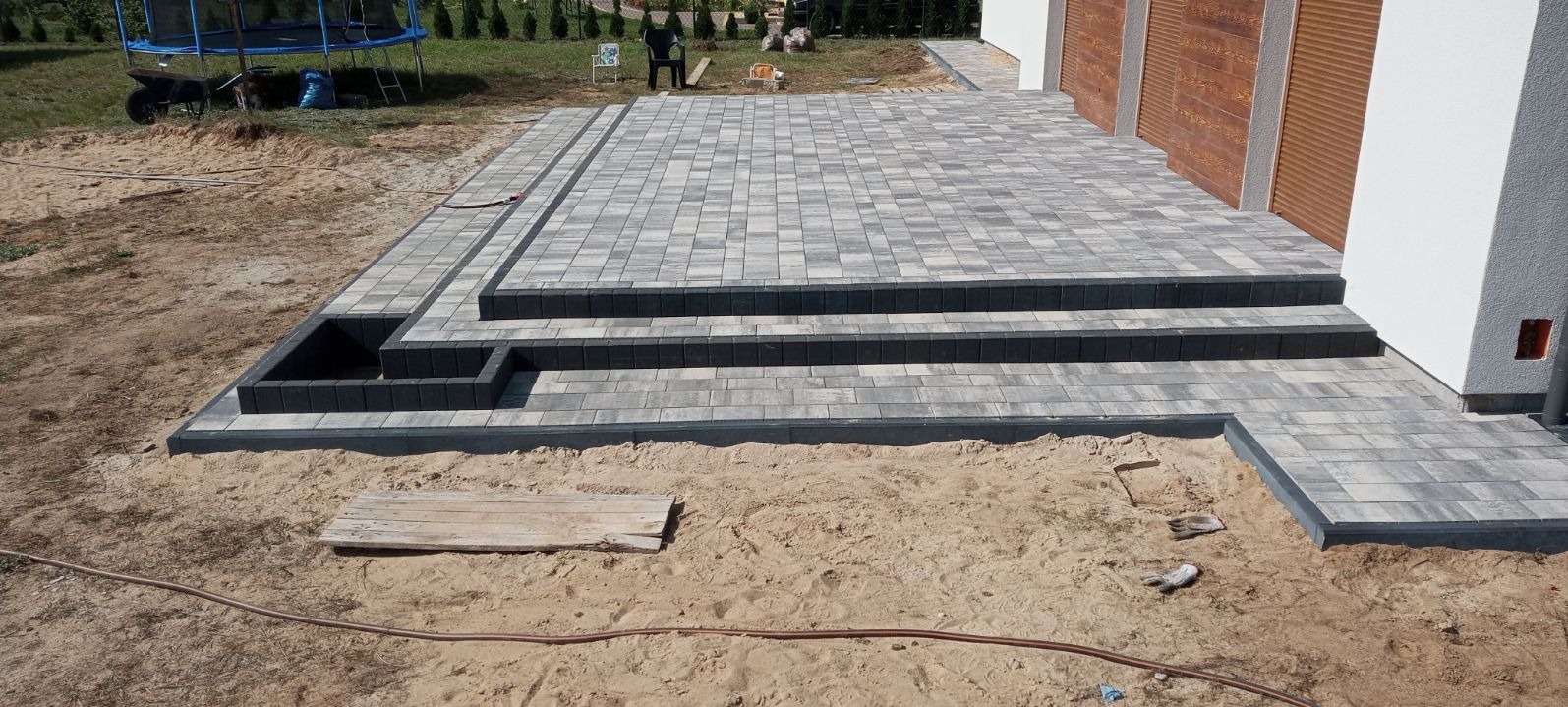 Ułożony taras z kostki brukowej z trzema stopniami, wykończony ciemnoszarym obrzeżem, widoczny piasek i narzędzia brukarskie w tle.