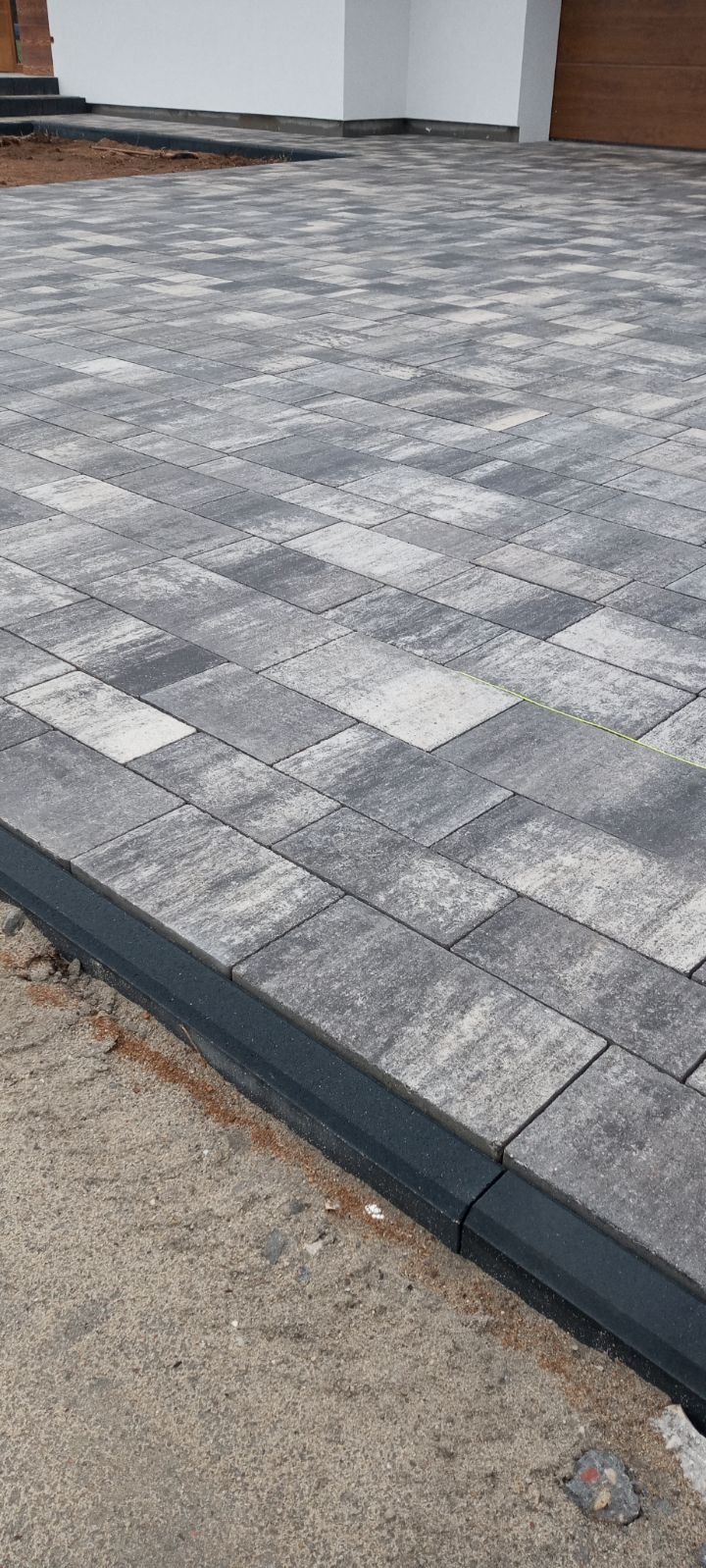 Ułożona, szara kostka brukowa o prostokątnym kształcie, z czarnym obrzeżem, na tle surowego terenu i fragmentu budynku.