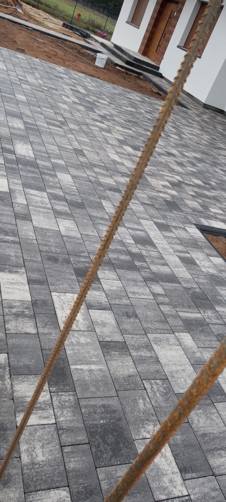 Ułożona szara kostka brukowa przed domem, z widocznym zbrojeniem w pierwszym planie i fragmentem budynku w tle.