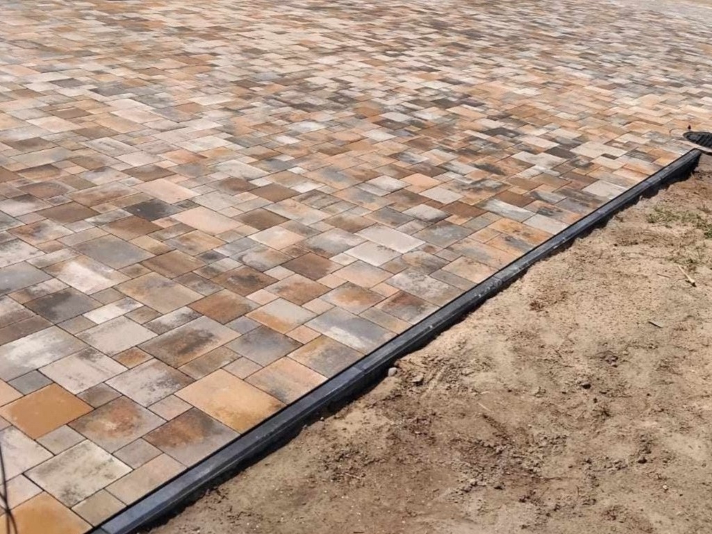 Ułożona nawierzchnia z kostki brukowej w odcieniach brązu i szarości, oddzielona czarnym obrzeżem od piaszczystego podłoża.