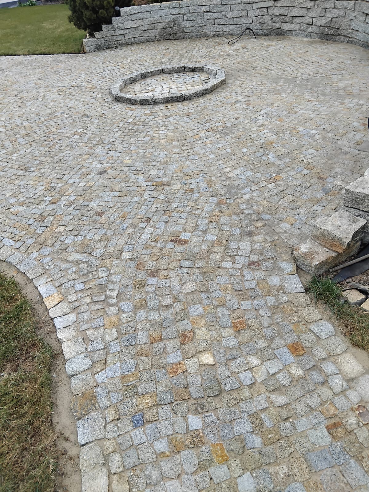 Ułożona z drobnej kostki granitowej nawierzchnia z okrągłym wzorem pośrodku, otoczona zielenią i elementami muru z kamienia.