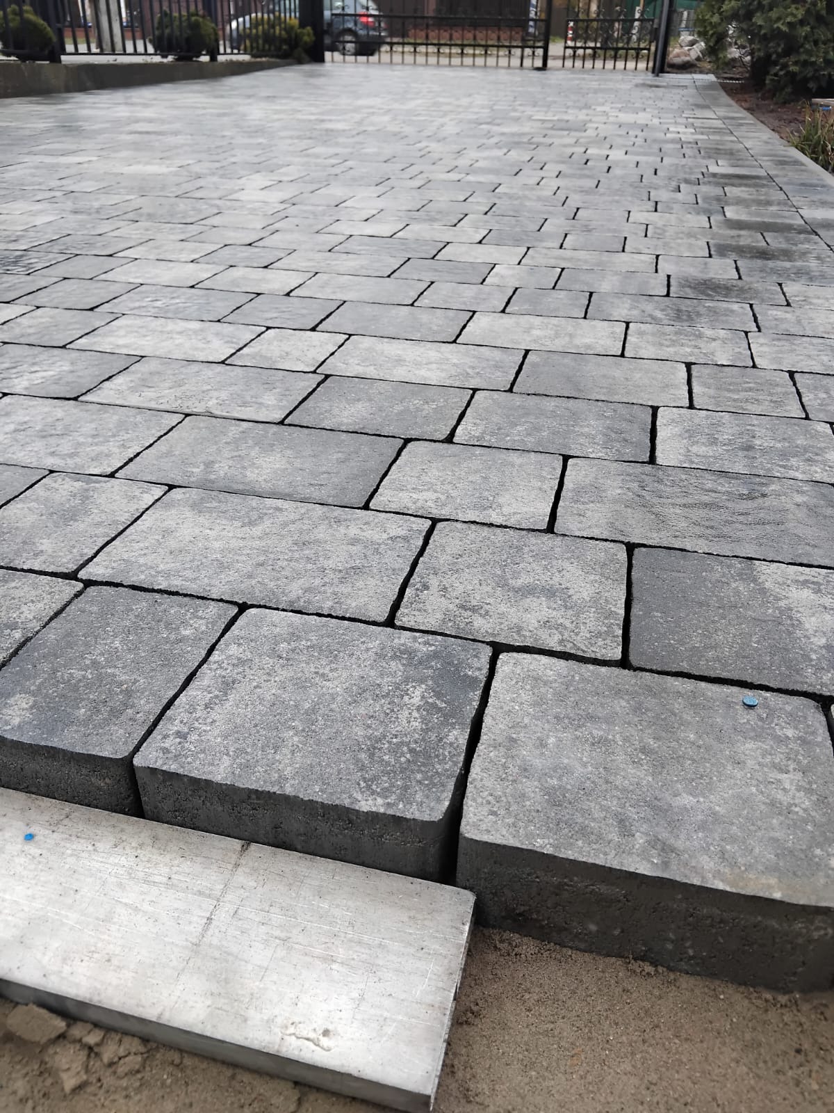 Ułożona szara kostka brukowa z widoczną linią cięcia i piaskiem w spoinach, w tle brama i zieleń, prezentująca efekt końcowy prac brukarskich.