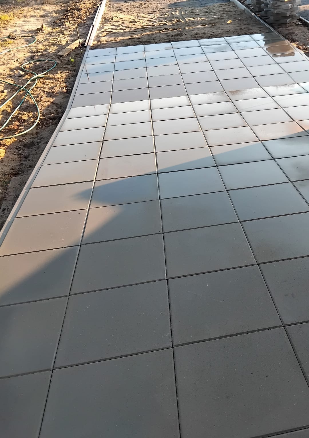 Świeżo ułożona, szara kostka brukowa w regularnym wzorze, delikatnie odbijająca światło, z widocznym przygotowanym terenem pod dalsze prace.