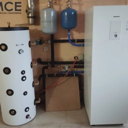 Małopolskie Centrum Ekologiczne Sp. z o.o. - Biały zasobnik Kospel, zbiorniki wyrównawcze i pompa ciepła Panasonic Aquarea, połączone rurami. Elementy instalacji grzewczej w pomieszczeniu.